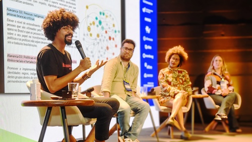 Samuel Emilio of Imaginable Futures sitting on stage and talking with a microphone.There are three more panelists on the stage with him. There is a backdrop with words and a chart.