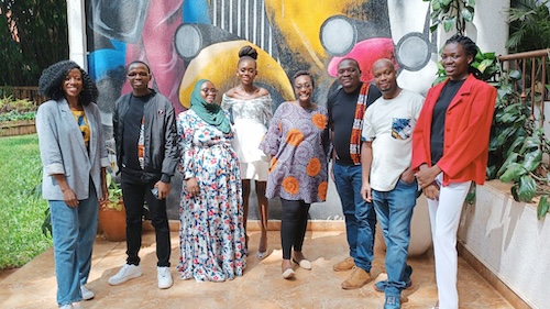 A group of 8 people stand together outdoors and are smiling. There is a colorful mural behind them.