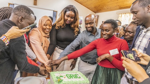 A group of people are smiling and cutting  a green cake together. The cake has the name "GRIC" on it.