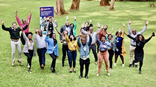 A group of 20 smiling people are standing on the grass outside with the arms up.  They look like they are cheering and are very excited. There is a banner that says "Imaginable Futures" in the back left corner. 