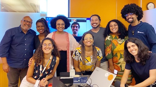 A group of 10 people in a meeting room smiling together at the camera. The walls in the room are orange.