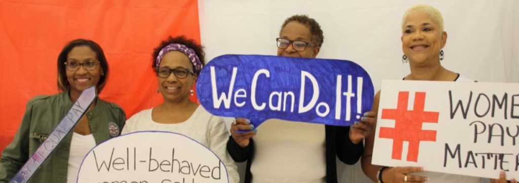 Three smiling women holding signs with women's empowerment messages that say: 1)"We Can Do it!" 2) "Equal pay" and 3) "Well-behaved women rarely make history."