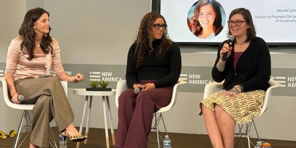 Three women sit onstage in a panel discussion, all smiling. The woman on the right is holding the microphone.