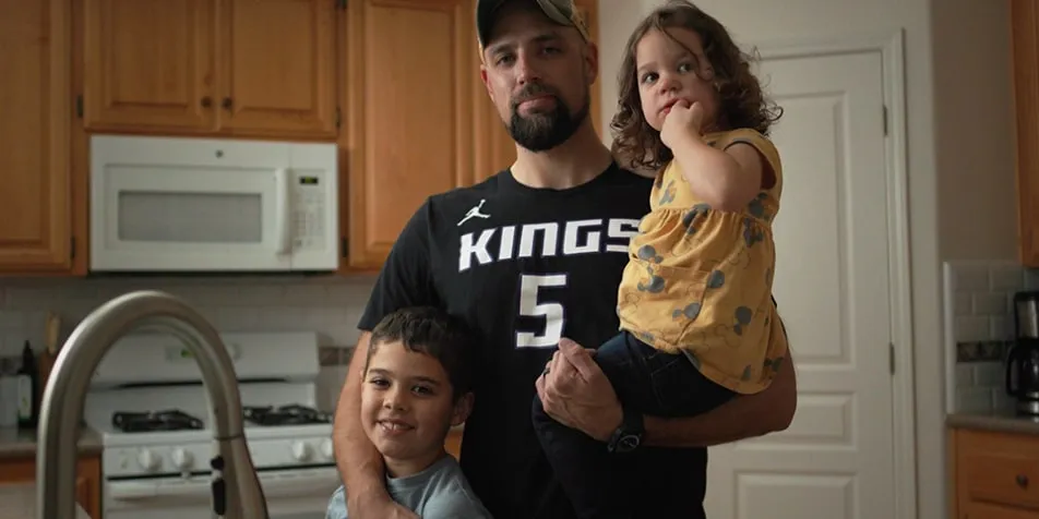 A bearded man wearing a Sacramento Kings basketball shirt stands in a kitchen, holding a young girl in a yellow shirt and standing beside a young boy in a gray shirt, all looking at the camera.