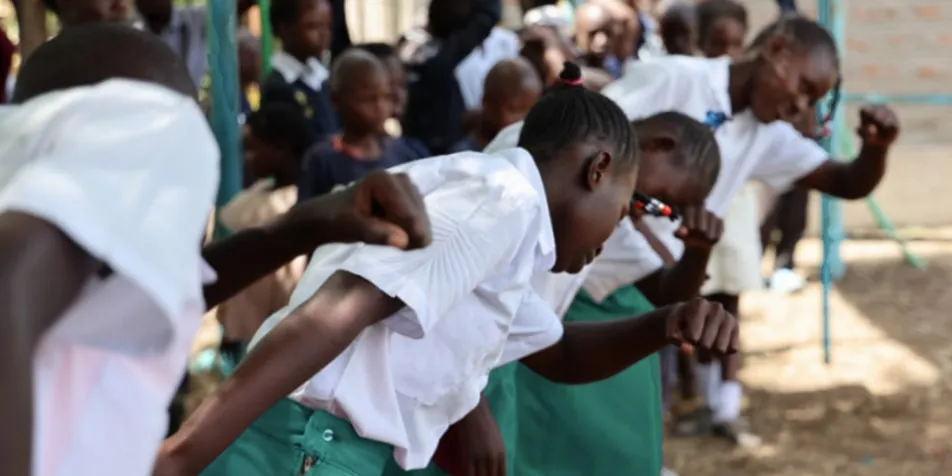 A group of children in school uniforms, including white shirts and green skirts, are actively participating in a physical activity outdoors, with other students and a swing set visible in the background.