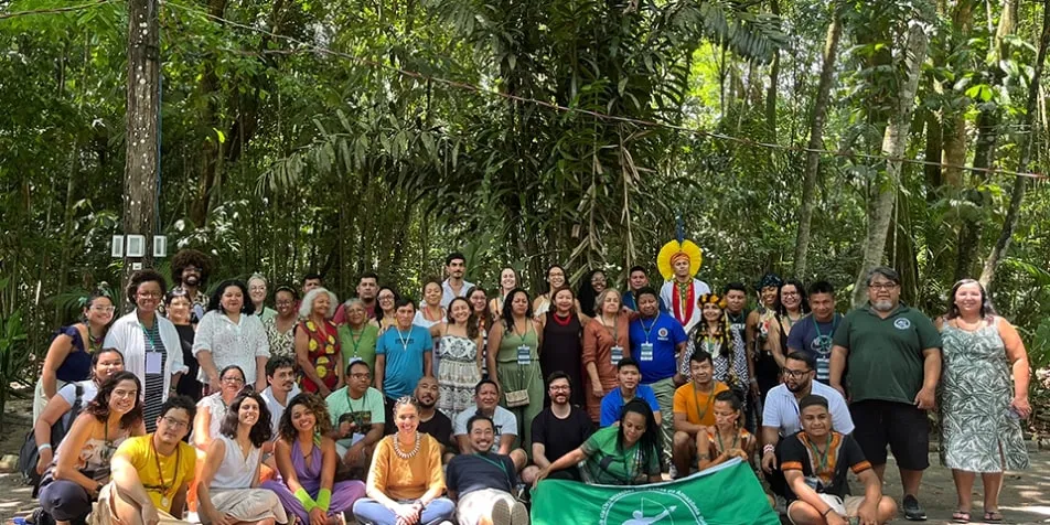 A group of people gathers outdoors in a forest area, posing for a group photo. Some hold a green banner at the front.