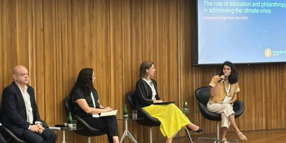 Four speakers sit on a panel in a wood-paneled conference room, discussing the role of education and philanthropy in addressing the climate crisis. A presentation screen with the topic is visible in the background.