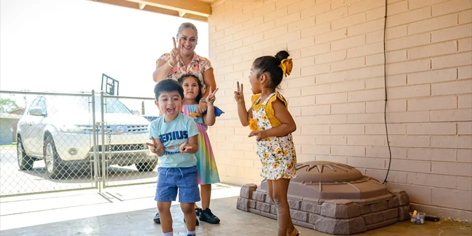 A caregiver plays with three young children on a covered outdoor patio. The children, two girls and one boy, are smiling and making peace signs with their hands, surrounded by chalk drawings on the concrete floor.