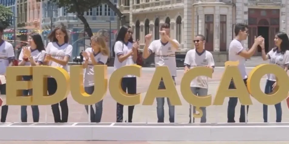 A group of people in white T-shirts standing outdoors, clapping and smiling behind large golden letters spelling "EDUCAÇÃO."