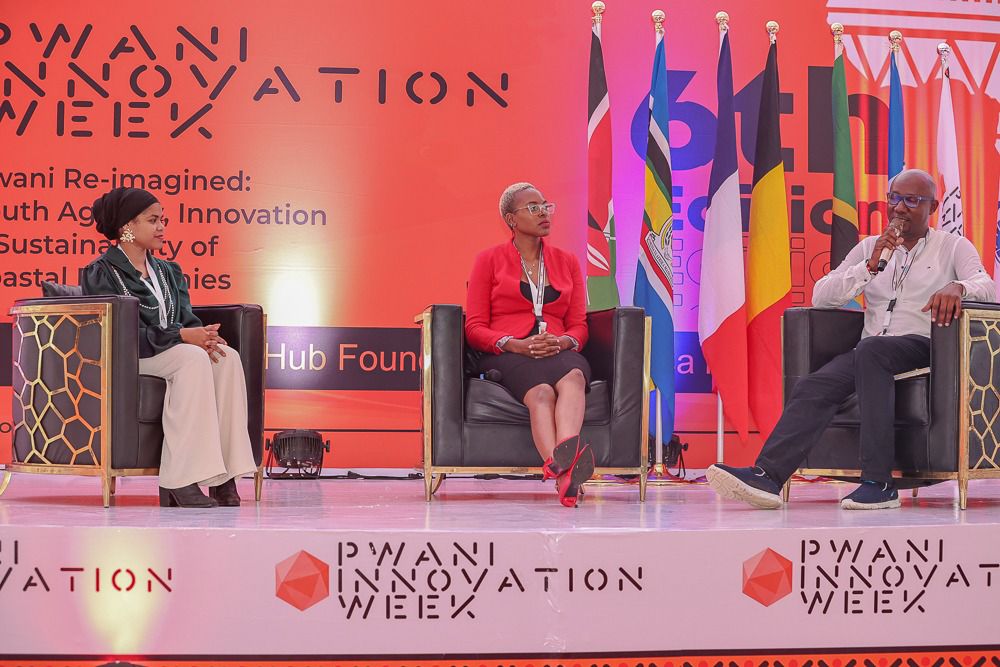 Três participantes do painel sentados no palco da Pwani Innovation Week, diante de um grande pano de fundo vermelho com a marca do evento e uma fileira de bandeiras internacionais. À esquerda, uma jovem com um lenço escuro na cabeça e blusa verde está sentada com as mãos cruzadas; no centro, uma mulher com blusa vermelha, saia preta e sapatos de salto vermelhos; à direita, um homem de camisa branca segurando um microfone. Na parte frontal do palco, está exibido o logotipo da Pwani Innovation Week.