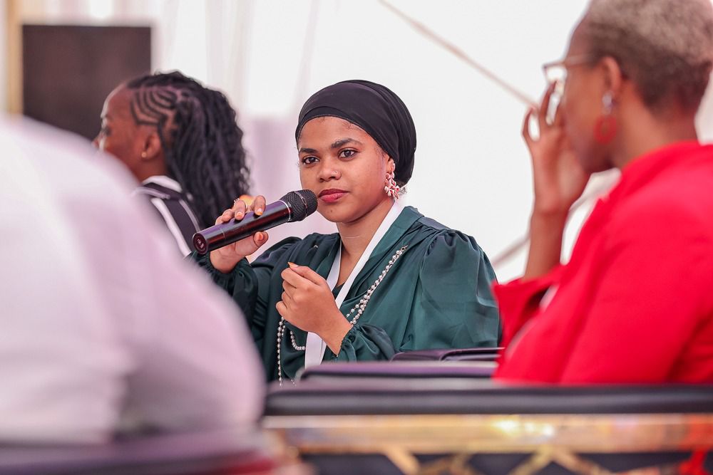A young woman in a black headwrap and dark green top speaks into a microphone, seated among other panel members under a marquee. Her expression is calm and focused.