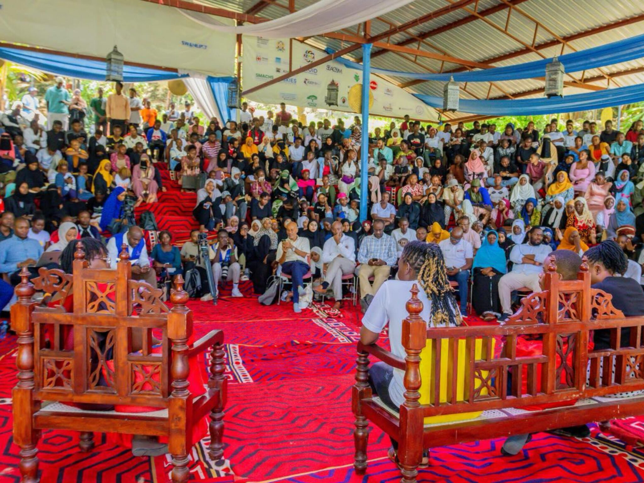Uma grande multidão de jovens sentados em um pavilhão aberto no Swahilipot Hub, ouvindo dois jovens palestrantes sentados em cadeiras de madeira esculpida e ornamentada em primeiro plano. Tapetes com padrões vermelhos cobrem o chão, tecidos drapeados em azul e branco pendem do teto, e uma faixa com as organizações parceiras se estende ao fundo do local.