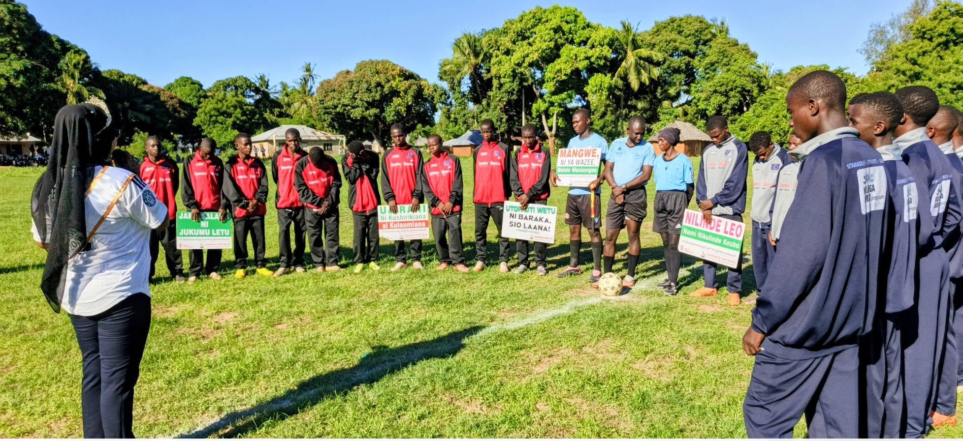 Duas equipes de futebol, vestidas com agasalhos esportivos, estão alinhadas em um campo gramado, com a cabeça baixa, enquanto árbitros em uniformes azuis, no centro, seguram uma bola de futebol. Vários jogadores seguram cartazes em suaíli promovendo a segurança da comunidade, a amizade e o respeito apesar das diferenças. Uma mulher com blusa branca e lenço preto na cabeça observa à esquerda, com palmeiras e casas comunitárias visíveis ao fundo.